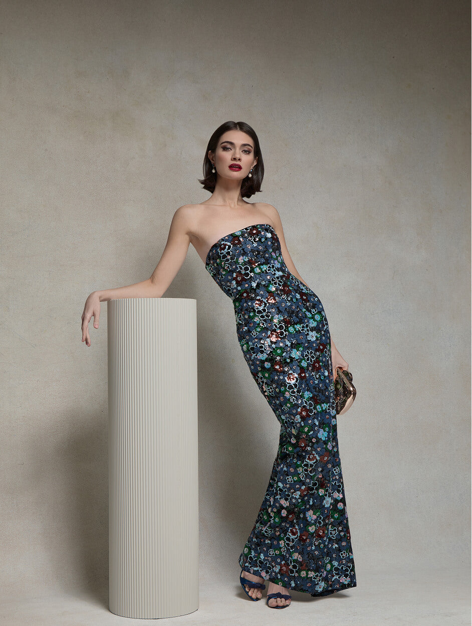 Model in a strapless blue and green floral column gown beside a cream pedestal.